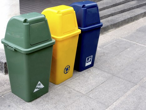 Three large, wheeled rubbish bins are positioned on a paved outdoor area near a building. The first bin, on the left, is green with a smooth, matte plastic surface and a sloped lid; it has a small triangular recycling symbol sticker near the bottom. The middle bin is bright yellow with a slightly textured plastic finish and features a black and yellow recycling icon on its side. The third bin, on the right, is dark blue with a matte plastic exterior and a slightly curved lid, displaying a small white symbol that appears to indicate general waste. All three bins have built-in handles or lids designed for easy access. They are situated directly on a grey stone pavement with visible joint lines, and the background includes a partial view of a building's steps or entrance area. The scene appears to be in an urban or commercial setting, possibly in Hanwell, in close proximity to a waste collection or rubbish disposal service, in relation to Office Clearance Hanwell's rubbish removal offerings.