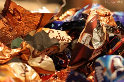 A close-up view of a pile of crumpled confectionery wrappers, primarily made of metallic foil and glossy paper with bright, reflective surfaces. The wrappers feature a mix of colors, including copper, silver, blue, and hints of red, and display various partial brand logos and text. The wrappers are irregularly crumpled, with some edges folded or twisted. Behind the pile, parts of other wrappers are visible, creating a cluttered and textured appearance typical of discarded sweet packaging. The setting appears to be indoors, possibly on a table or tray, with soft ambient lighting highlighting the shiny surfaces. This image visually represents the aftermath of confectionery consumption, relevant to rubbish or waste associated with confectionery packaging, which would be handled by a rubbish removal service like Office Clearance Hanwell in the local area of Hanwell postcodes.