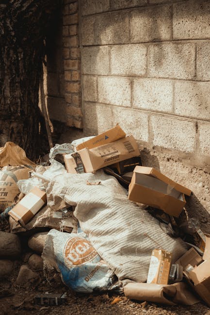 A collection of discarded cardboard boxes, some flattened and others partially assembled, piled against a rough concrete and brick wall in an outdoor setting. The boxes vary in size, with brown corrug