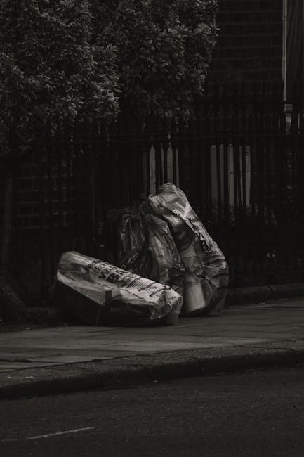 A collection of discarded cardboard boxes, some flattened and others partially assembled, piled against a rough concrete and brick wall in an outdoor setting. The boxes vary in size, with brown corrug