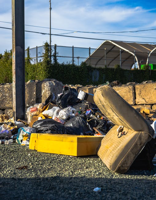 A large pile of mixed waste materials is scattered on a gravel surface adjacent to a low stone wall. The waste includes black and transparent plastic bags, some torn open, revealing assorted household