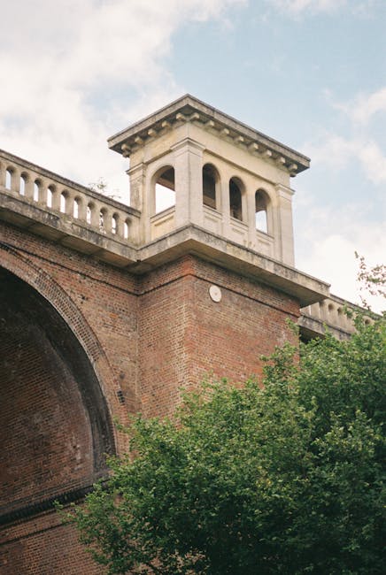 A large stone viaduct with multiple semi-circular arches spans a deep, green valley filled with dense trees and foliage. The viaduct's weathered grey surface contrasts with the rich, leafy textures of