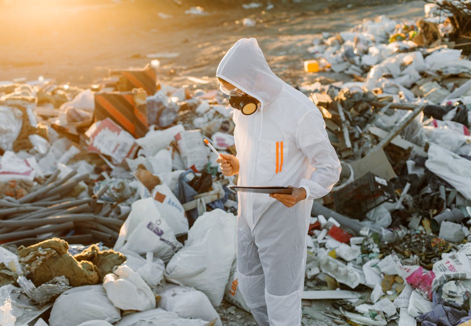 A large accumulation of mixed waste and debris is scattered along the shoreline of a small body of water, with a steep, eroded dirt bank in the background. The waste includes various plastic bags, bot